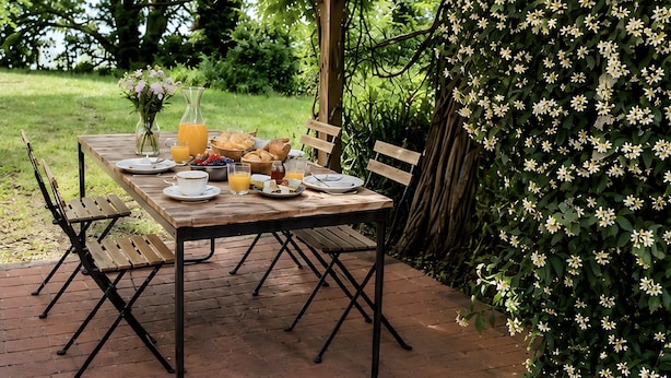 Un espace de restauration en plein air avec une table dressée pour le petit-déjeuner, entouré de verdure et d’un arbuste en fleurs.