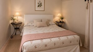 Une chambre à coucher avec un lit, deux tables de chevet, une estampe botanique encadrée et une fenêtre avec des rideaux.