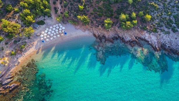 Vista aerea di una spiaggia con acque turchesi cristalline, una costa rocciosa e una fila di ombrelloni bianchi.