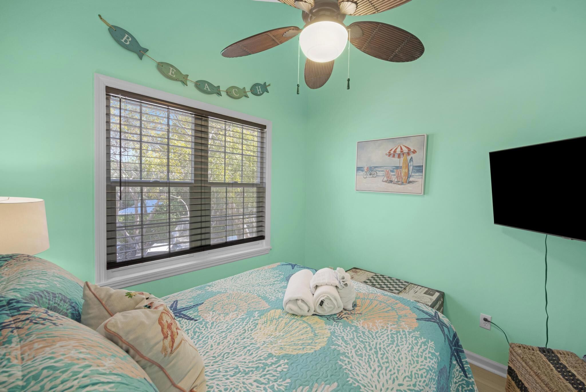 Une chambre à coucher comprenant un lit, un ventilateur de plafond, une fenêtre avec des stores, un téléviseur fixé au mur et un tableau encadré.