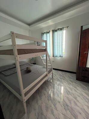 A bunk bed with a mattress and pillows, a small table with a chair, a window with curtains, and a door. - Yena's Inn (Cauayan)