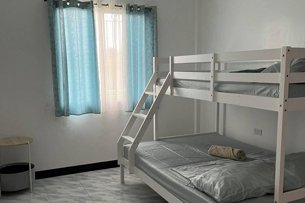 A bunk bed room with a window, curtains, a small table, and a trash bin.