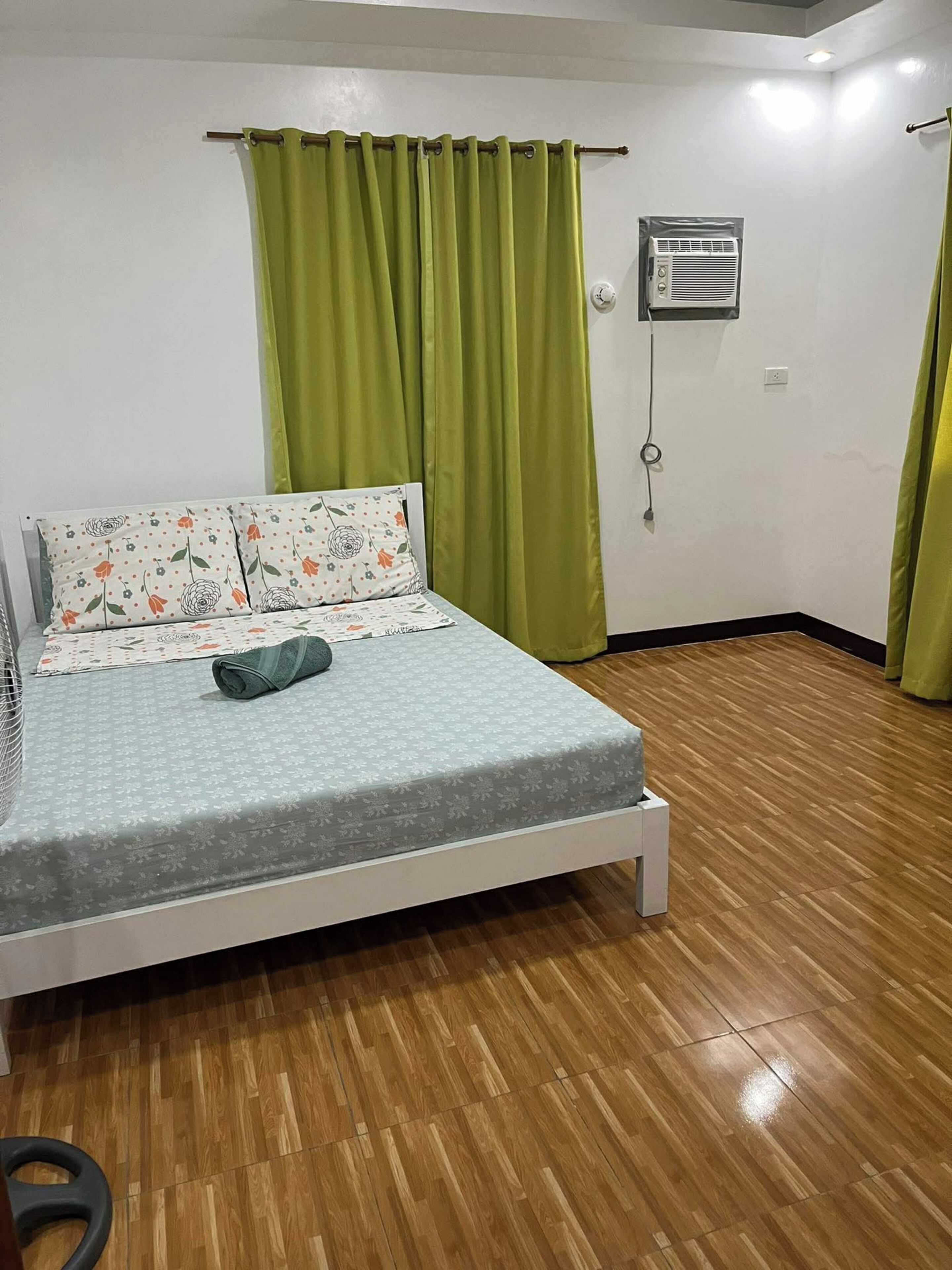 Ein Einzelbett mit einem blumengemusterten Kopfkissen, ein grüner Vorhang und eine Wand-Klimaanlage in einem Raum mit Holzboden.