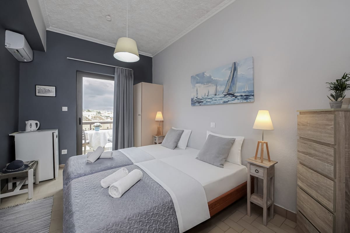 Habitación de hotel con cama, lámparas de noche, un cuadro, un escritorio con hervidor y vistas a través de una puerta.