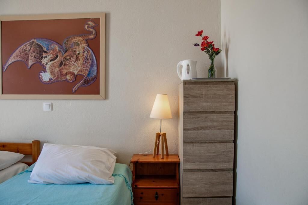 A bedroom with a bed, a nightstand with a lamp, a wooden dresser, a framed artwork, and a vase with flowers.