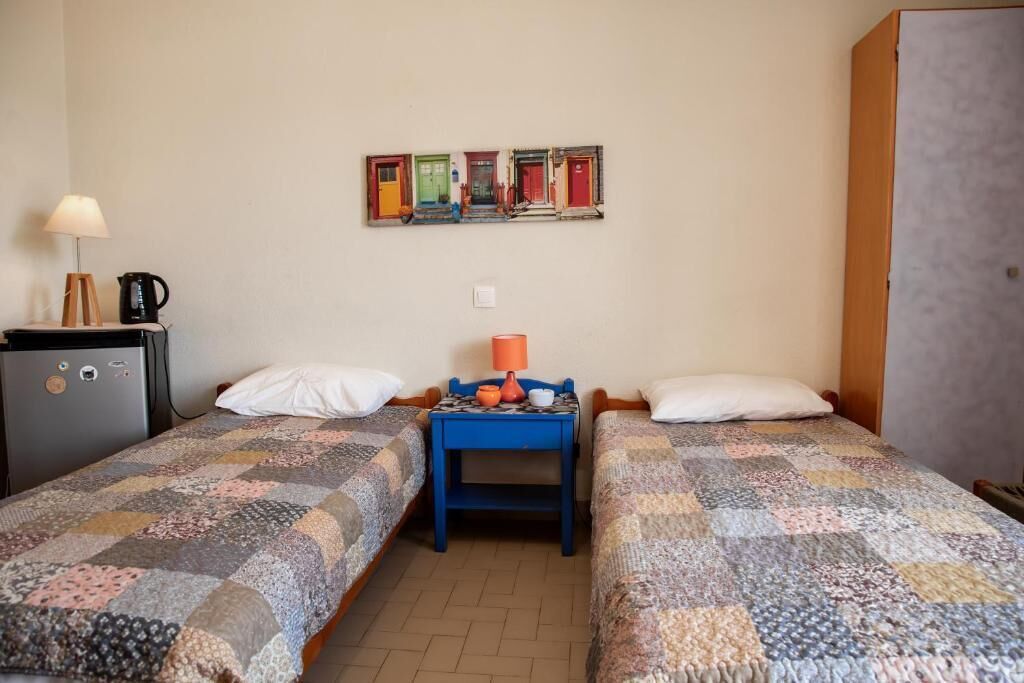A room with two beds, a small table with a lamp, a refrigerator, and a wall-mounted artwork.