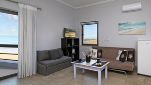 Un salón con un sofá gris, un sofá marrón, una pequeña mesa con adornos, una estantería negra, una ventana con vista a la playa y un equipo de aire acondicionado.