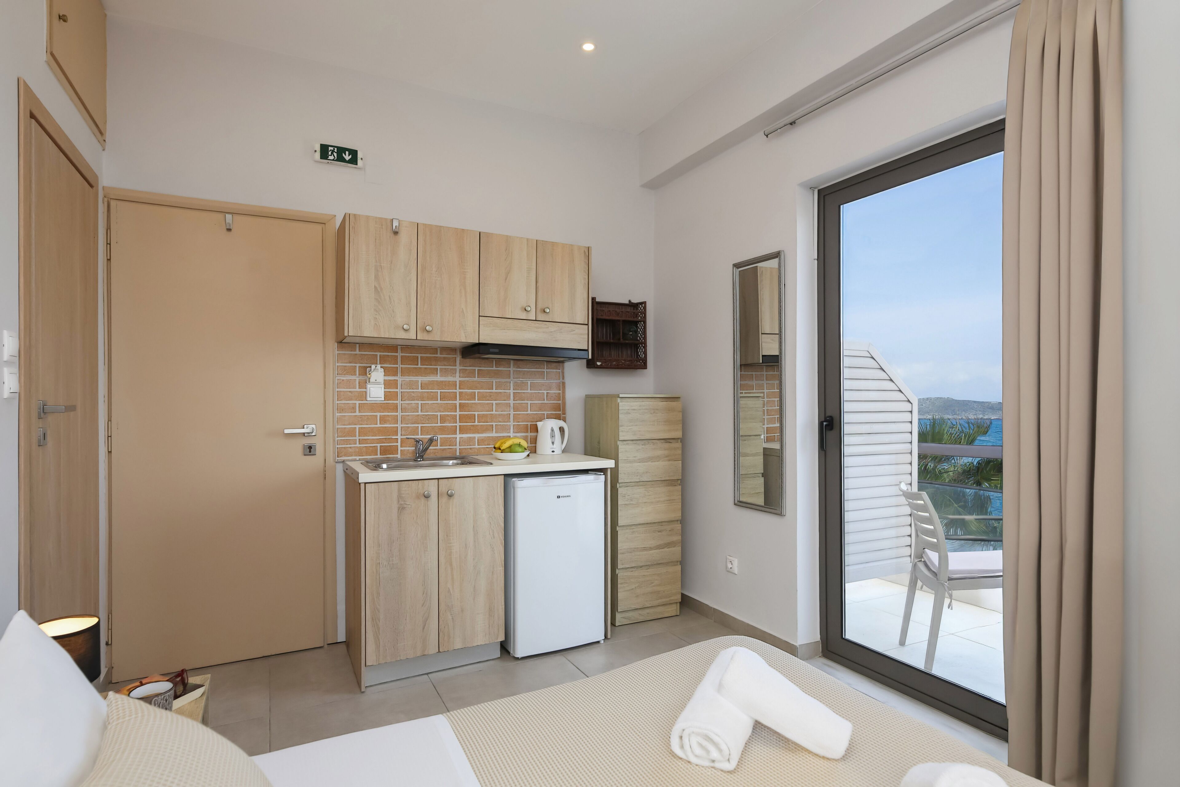 A compact hotel room with a kitchenette, a balcony with a chair, and a view of the outdoors.