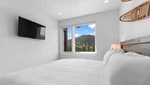A bedroom with a bed, a TV mounted on the wall, a window with a mountain view, and a wicker chair. - Luxury Keystone Condo - Lodge & Spa Access & 5 Min Drive to Slopes! (Keystone)