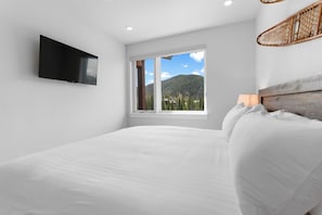A bedroom with a bed, a TV mounted on the wall, a window with a mountain view, and a wicker chair. - Luxury Keystone Condo - Lodge & Spa Access & 5 Min Drive to Slopes! (Keystone)