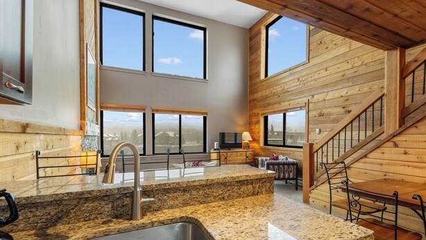 Una cocina con encimeras de granito, pileta de acero inoxidable, paredes de madera y ventanas amplias.