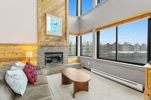 A living room with a stone fireplace, a large window with a view of buildings, a sofa with cushions, and a wooden coffee table. - Lake-View Condo - 5 Min Drive to Keystone Ski Village (Keystone)
