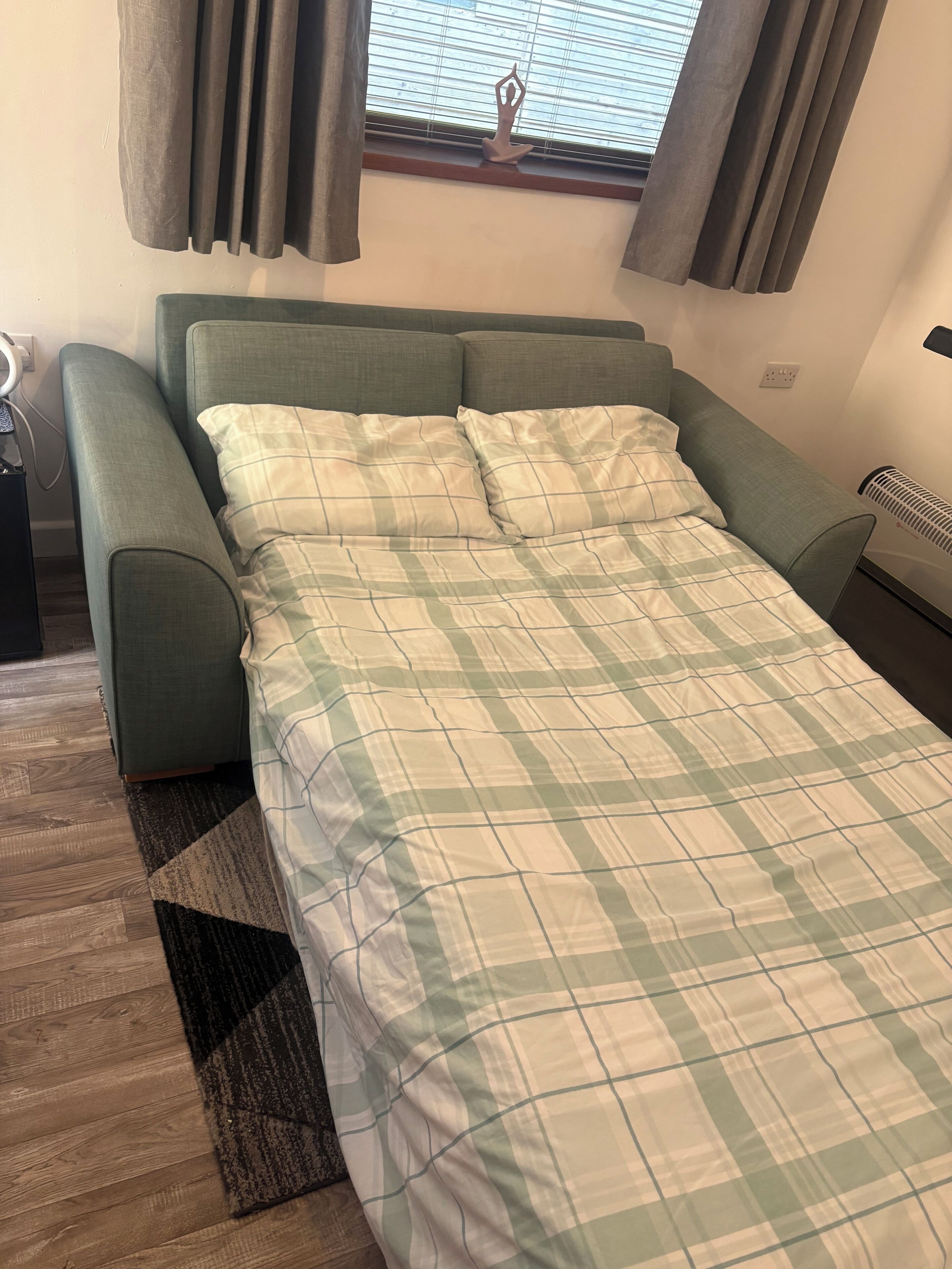 Una cama individual con colcha escocesa, un sofá tapizado en verde, una ventana con persianas y un radiador.