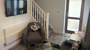 Un salon avec un fauteuil gris, un coussin décoratif à motifs, un téléviseur à écran plat fixé au mur, un escalier en bois et une porte avec un panneau de verre.