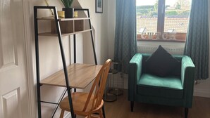 Une pièce avec un plancher en bois, un bureau avec une chaise, un canapé vert, un pouf noir, une plante en pot, un tableau encadré et une fenêtre avec des rideaux.