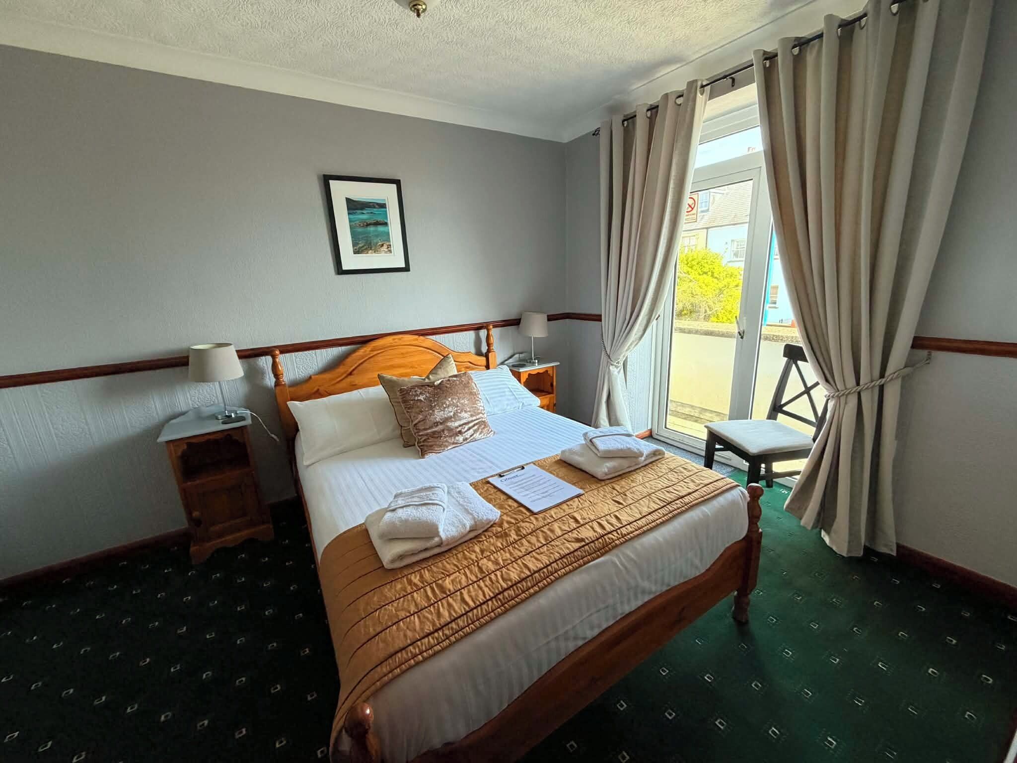 Ein Hotelzimmer mit einem Bett, einem Nachttisch, einer Lampe, einem gerahmten Bild an der Wand und Blick ins Freie durch das Fenster.