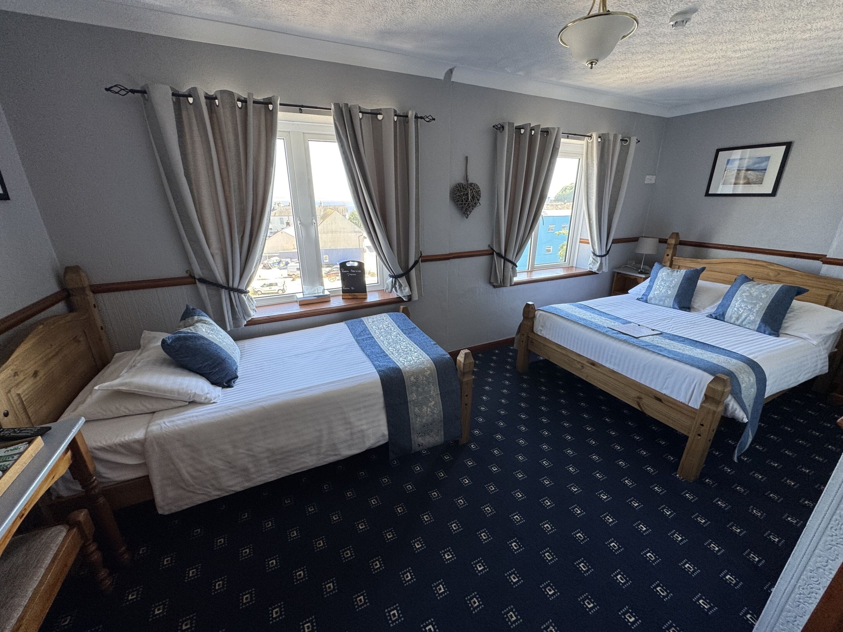 A room with two wooden beds, a window with curtains, a small table, and a wall-mounted decoration.