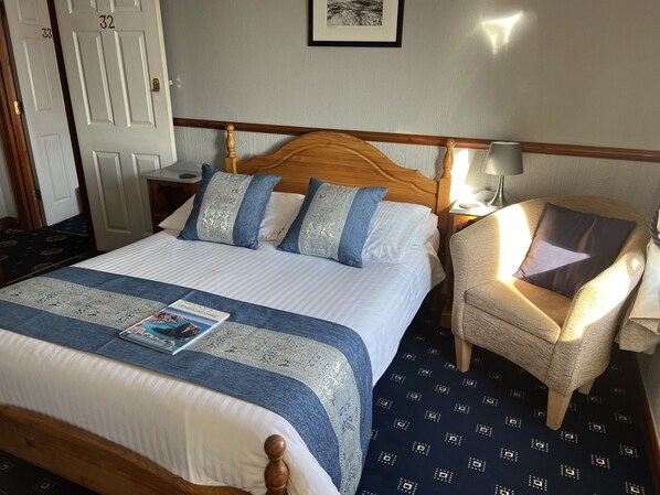 A neatly made bed with a blue and white bedspread, a wooden headboard, and a chair with a cushion. - The Gower Hotel (Saundersfoot)