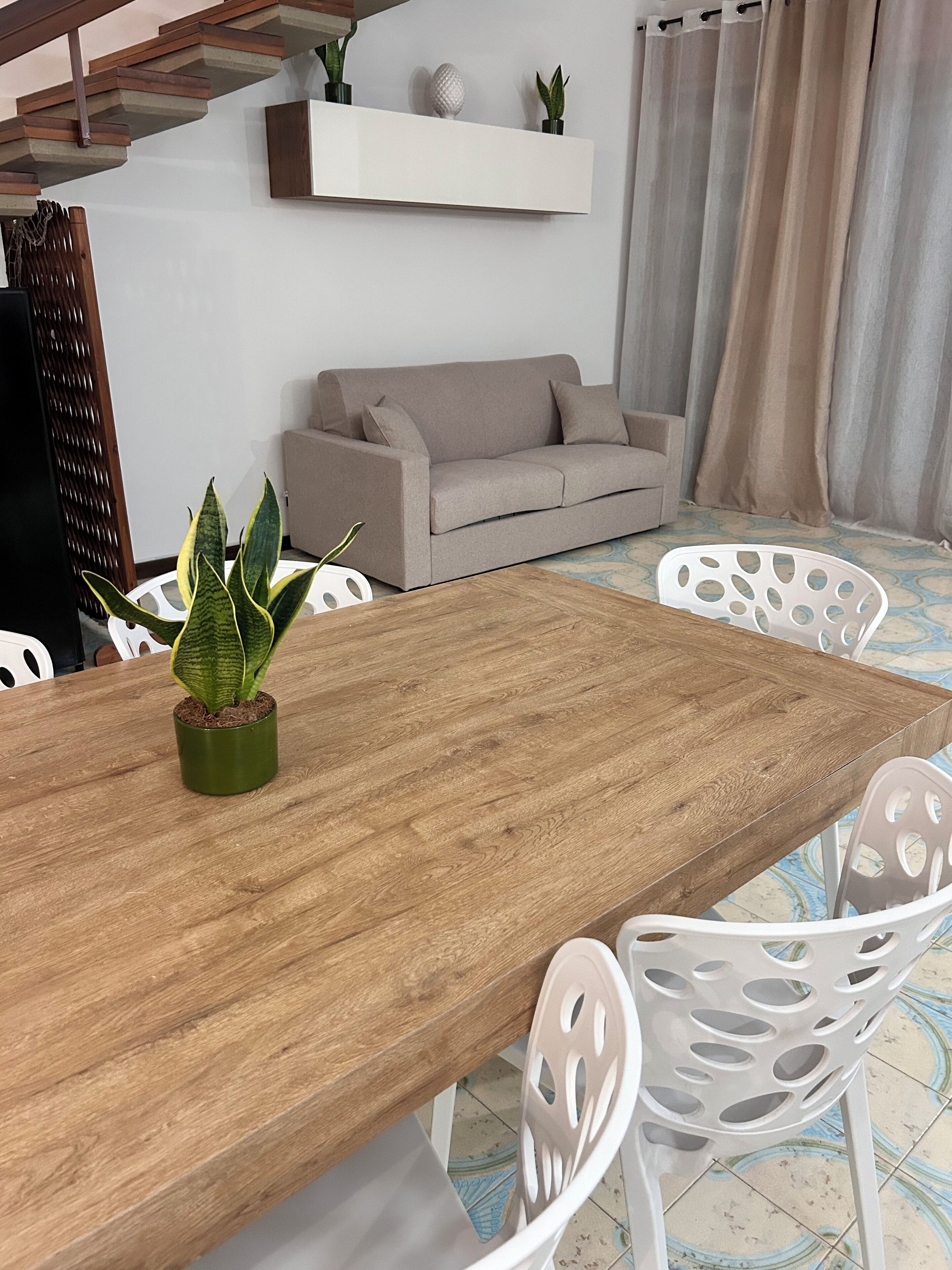 A dining area with a wooden table, white chairs, a grey sofa, and a potted plant.