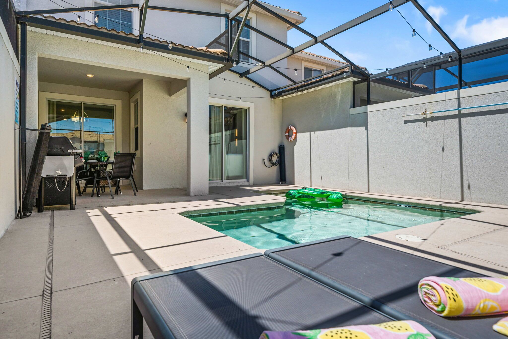 Uma piscina com espreguiçadeiras, uma mesa e cadeiras, e uma área de varanda envidraçada.