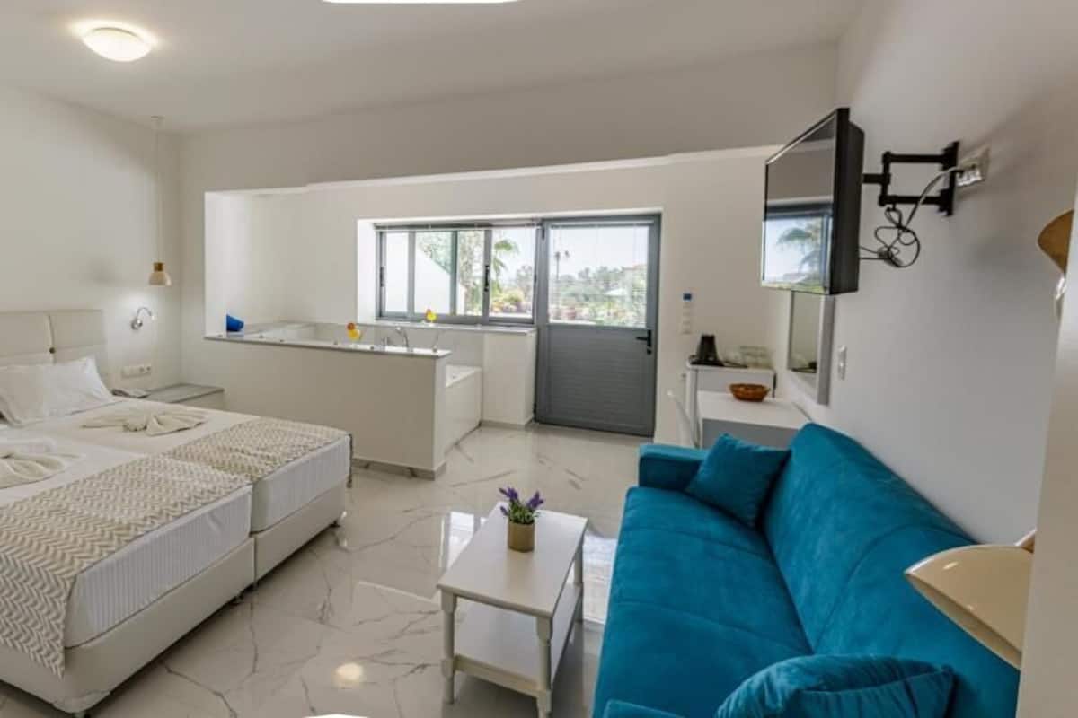 A modern hotel room with two beds, a blue sofa, a small table, a kitchen area, and a large window.