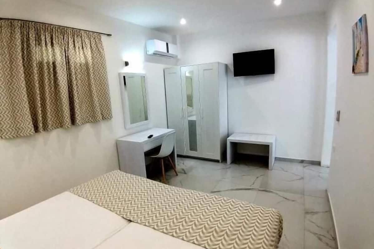 A bedroom with a bed, a desk, a chair, a wardrobe, a television, and a window with curtains.