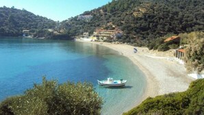 Escena costera con un barco en agua tranquila, una playa arenosa y edificios en una colina.