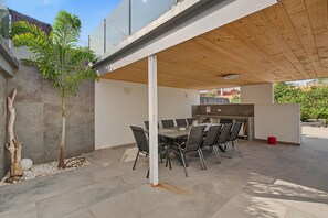 An outdoor patio with a dining table and chairs, a small tree, and a kitchen area.
