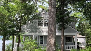 A two-story house with a porch, surrounded by trees and a body of water. - Classic NH Lake House | Sandy Beach, 2 Boat Dock, Sleeps 10 (Mirror Lake)