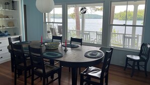 A dining area with a round table, chairs, a hanging light fixture, and large windows overlooking a body of water. - Classic NH Lake House | Sandy Beach, 2 Boat Dock, Sleeps 10 (Mirror Lake)