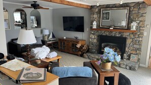 A living room with a stone fireplace, a flat-screen TV, and a ceiling fan. - Classic NH Lake House | Sandy Beach, 2 Boat Dock, Sleeps 10 (Mirror Lake)