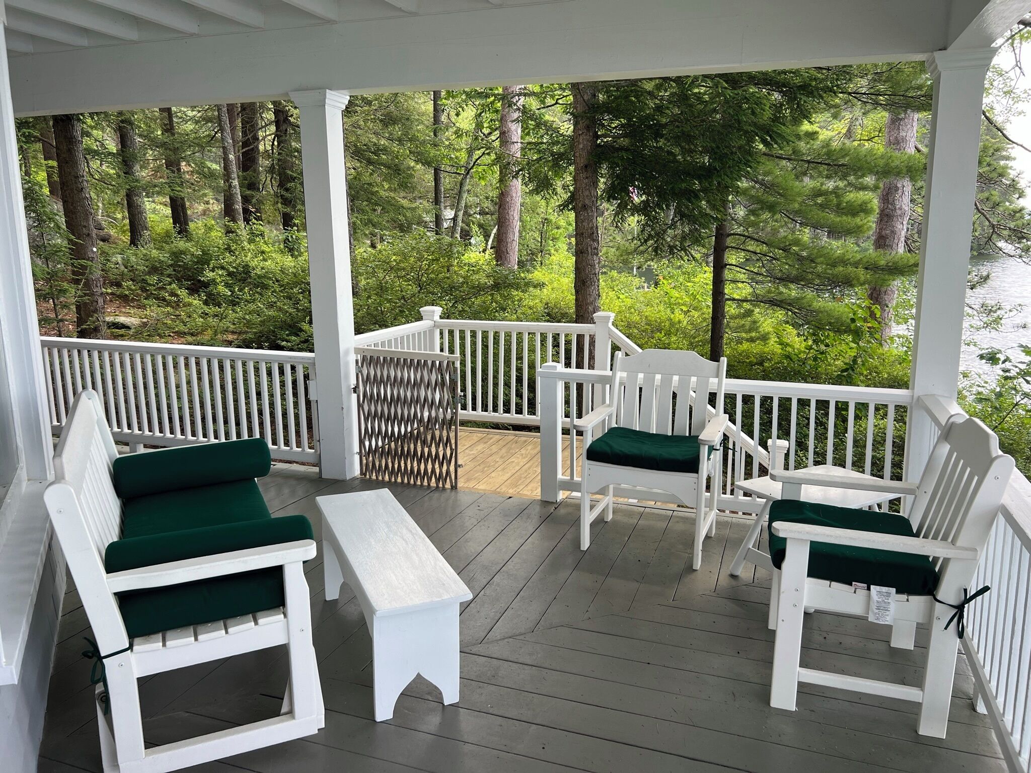 A covered porch with white wooden furniture and green cushions, surrounded by a white railing, overlooking a forested area.