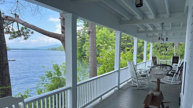 A covered porch with white railings, rocking chairs, and a table. - Classic NH Lake House | Sandy Beach, 2 Boat Dock, Sleeps 10 (Mirror Lake)