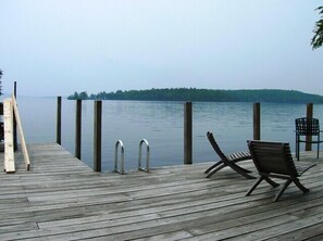 Une jetée en bois, dotée de deux échelles métalliques et de deux chaises, fait face à un lac paisible, avec une île boisée visible au loin.