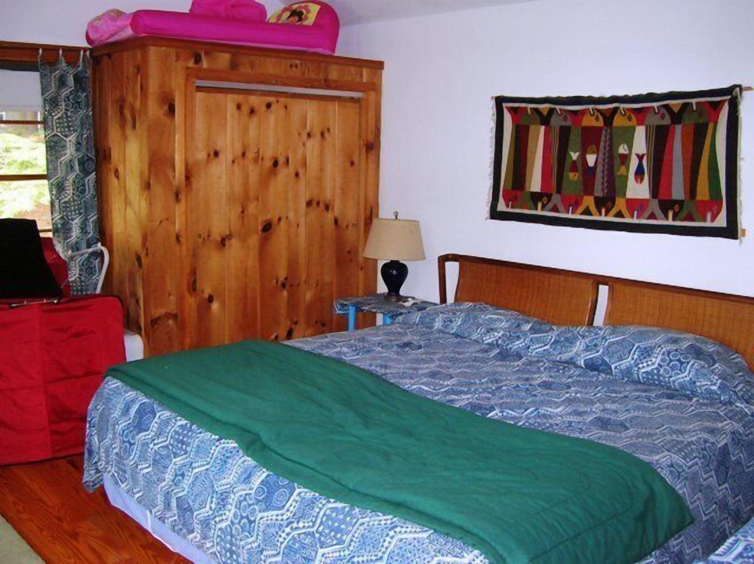 Una habitación con una cama, un armario de madera, una silla roja, una mesita con una lámpara y un tapiz de pared colorido.
