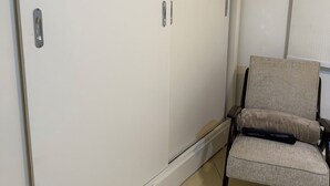 Huoneessa on suuri valkoinen vaatekaappi, harmaa verhoiltu tuoli ja laatoitettu lattia.