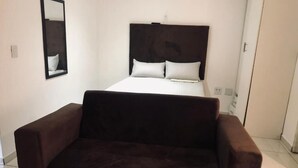 A room with a bed, a dark brown sofa, a wall-mounted mirror, and a door. - UNCLE CHARLES GUESTHOUSE (Pretoria)