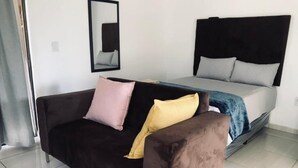 A room with a bed, a dark brown sofa with two pillows, a mirror on the wall, and a red patterned rug. - UNCLE CHARLES GUESTHOUSE (Pretoria)