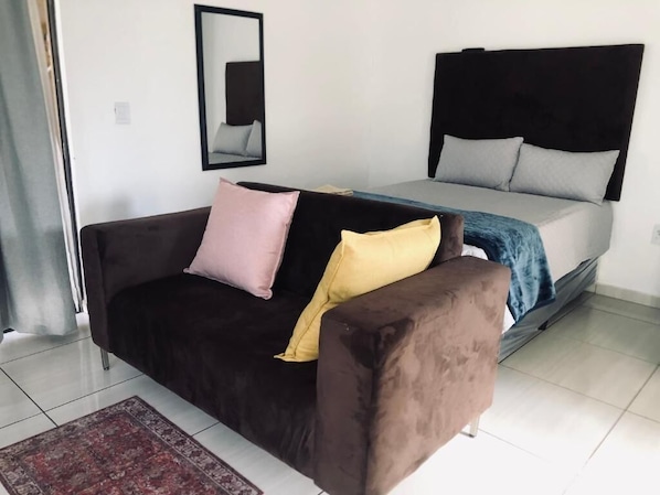 A room with a bed, a dark brown sofa with two pillows, a mirror on the wall, and a red patterned rug. - UNCLE CHARLES GUESTHOUSE (Pretoria)