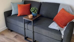 A grey sofa with orange and blue cushions, a small wooden side table with a plant and bowls, and three paintings on the wall. - Cozy Cottage - Lakeview (Reeds Spring)