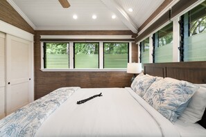 A bedroom with a large bed, a wooden headboard, a window with a view of a green field, and a ceiling fan. - Spacious 2BD  2BA Cottage Pools Walk to North Kaanapali Beach - ICC 2 (Lahaina)