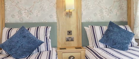 A hotel room with two beds, each with striped bedding and blue pillows, a wooden nightstand with drawers, and a wall-mounted light fixture.