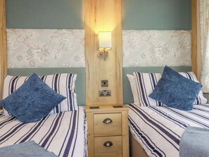 A hotel room with two beds, each with striped bedding and blue pillows, a wooden nightstand with drawers, and a wall-mounted light fixture. - 8 Summerleaze (Bude)