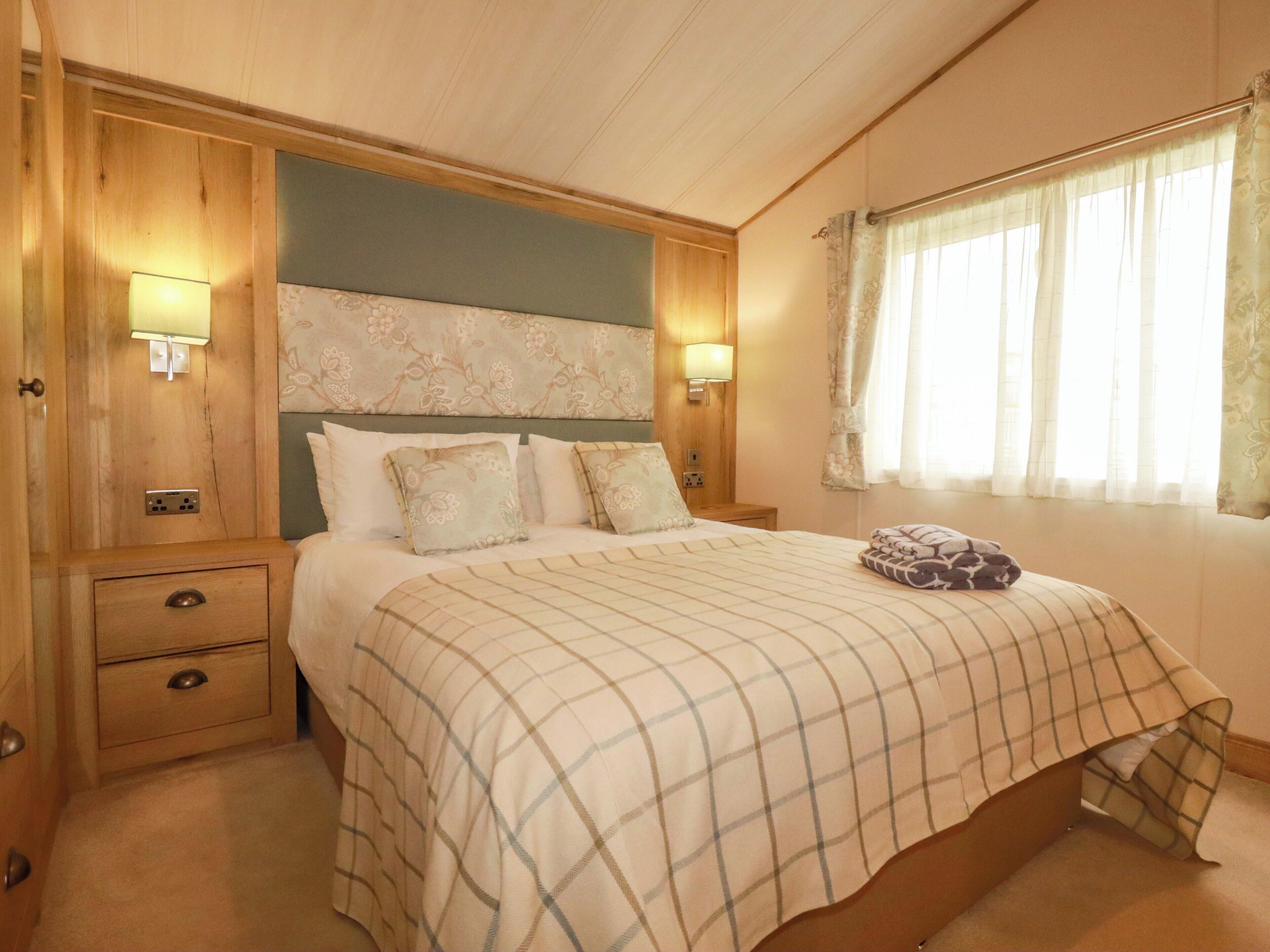 Un dormitorio con cama, lámparas de noche, mesita de noche, cajón y ventana con cortinas.