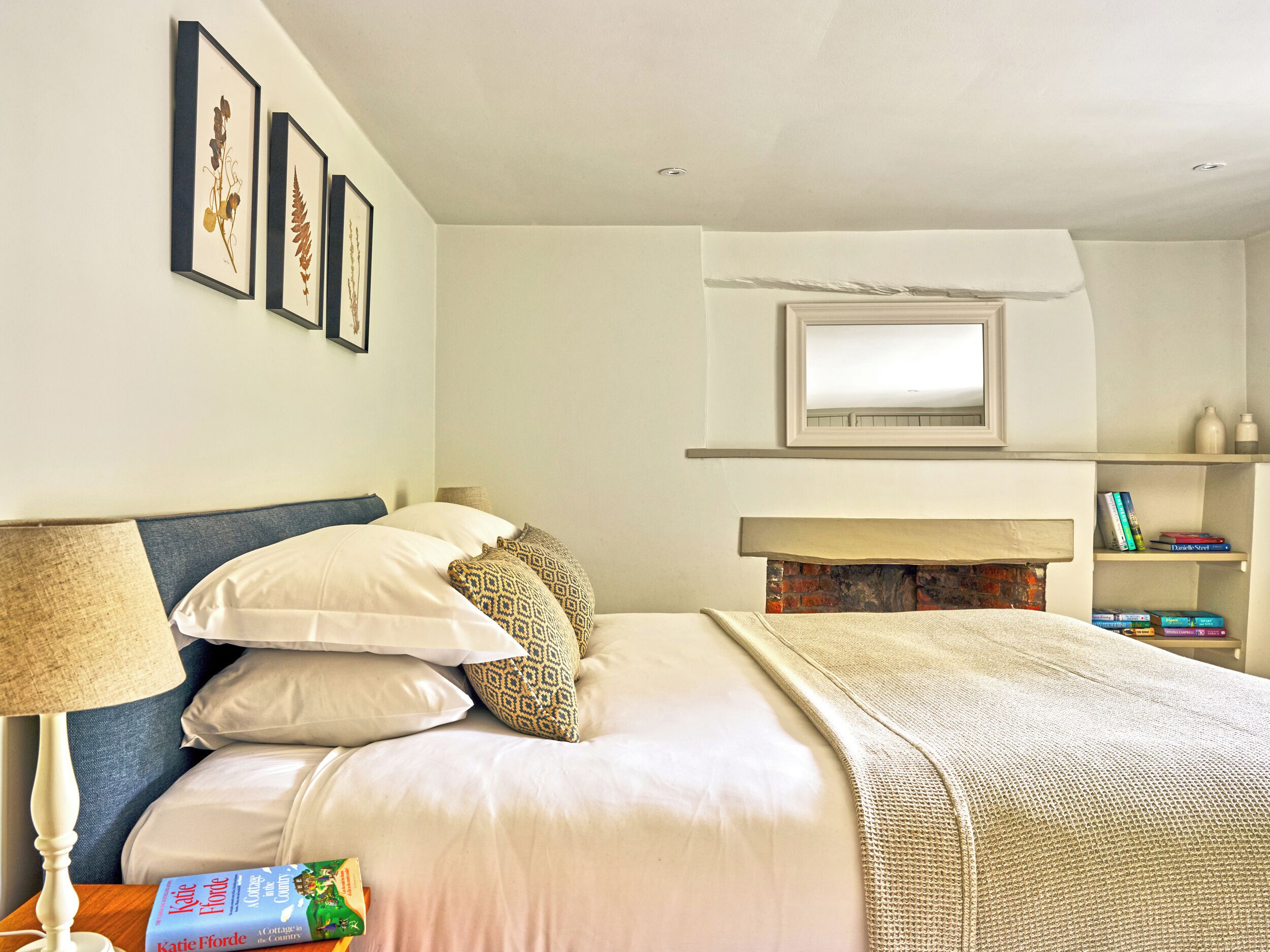 A neatly arranged bedroom with a bed, pillows, a bedside lamp, a fireplace, framed pictures, and a bookshelf.