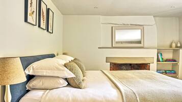 A neatly arranged bedroom with a bed, pillows, a bedside lamp, a fireplace, framed pictures, and a bookshelf.