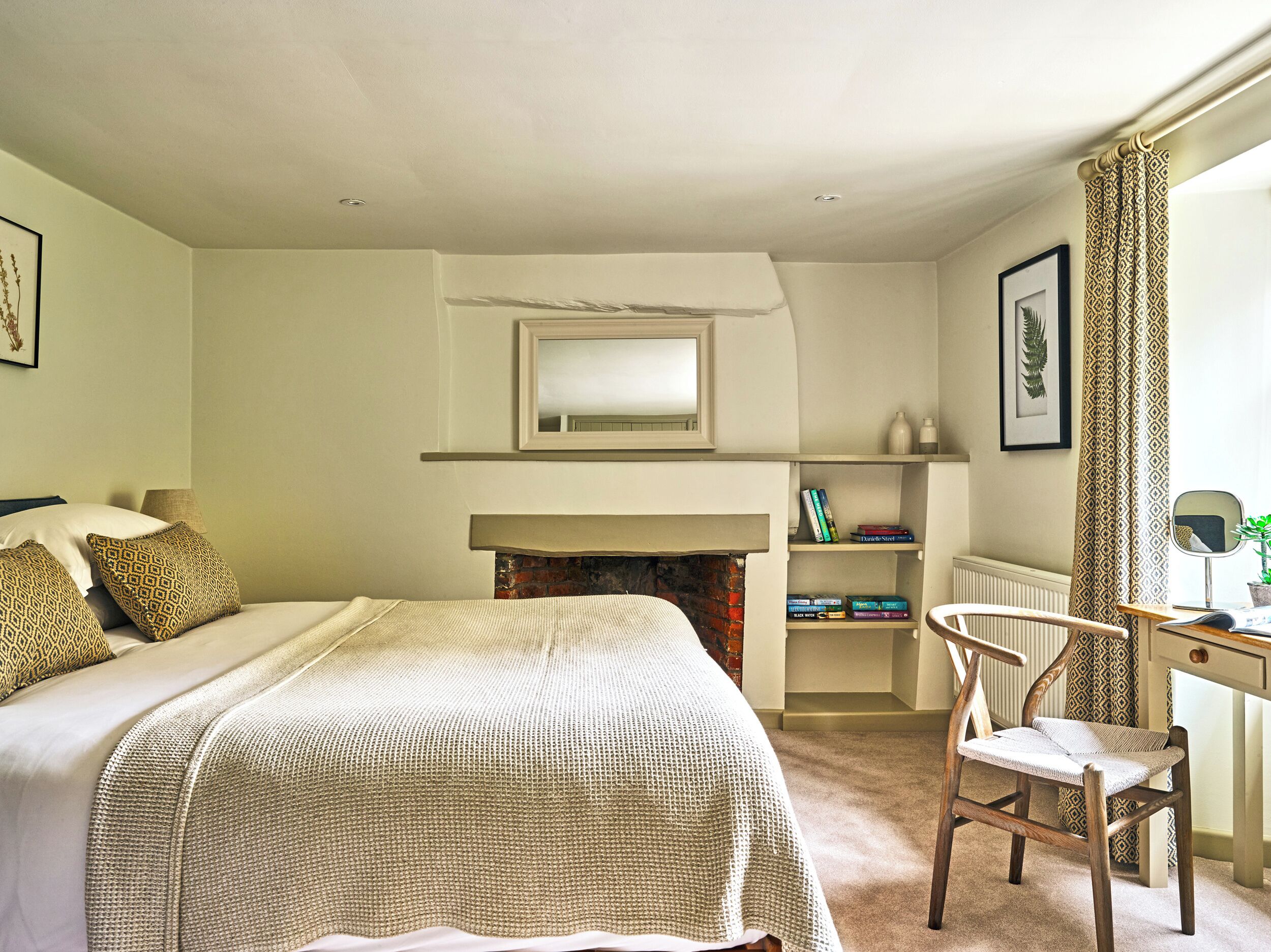 A bedroom with a bed, a chair, a desk, a fireplace, and a mirror.