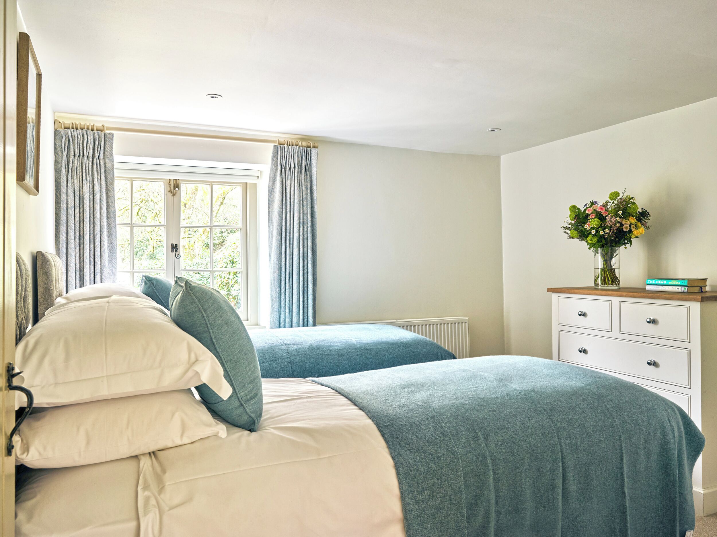 Un dormitorio con dos camas, un aparador, una ventana con cortinas y un florero con flores.