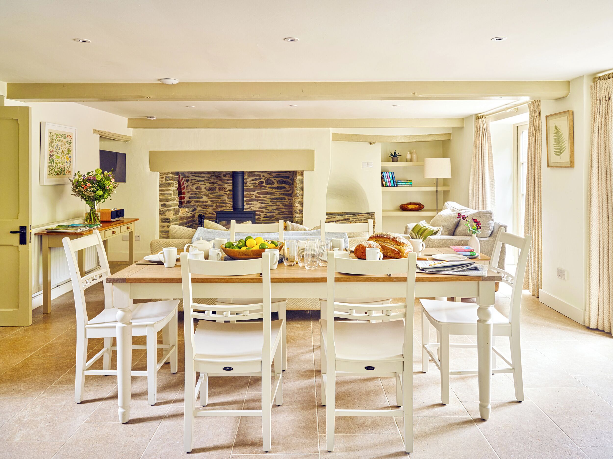A dining room with a large table, white chairs, a stone wall, and a cozy living area with a sofa and a lamp.