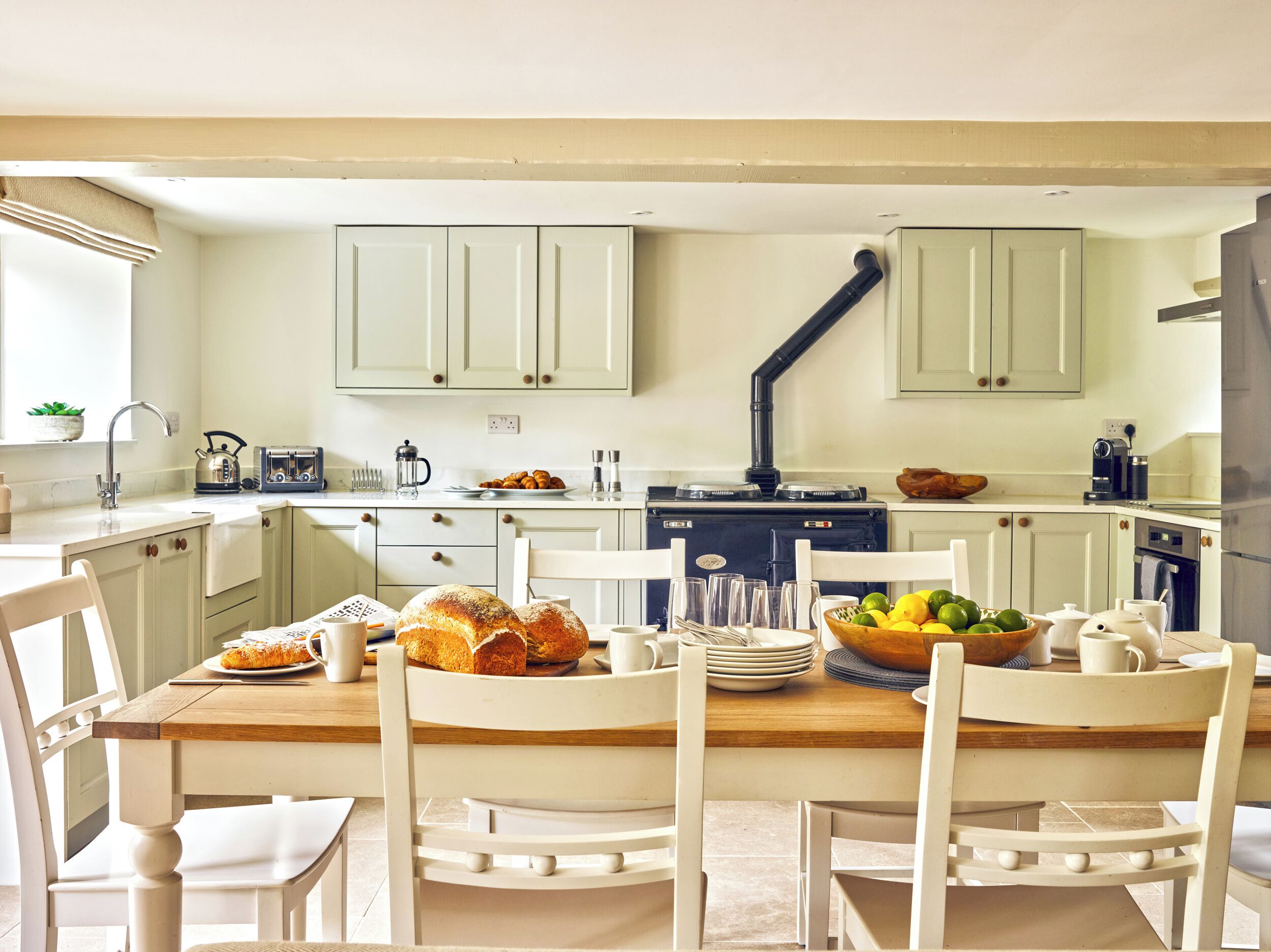 A kitchen with white cabinets, a wooden dining table, and chairs. There is a stove, a sink, and various kitchen items on the countertops.
